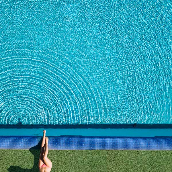 Fotografía de una piscina vista desde arriba con las piernas de una chica joven tomando el sol.